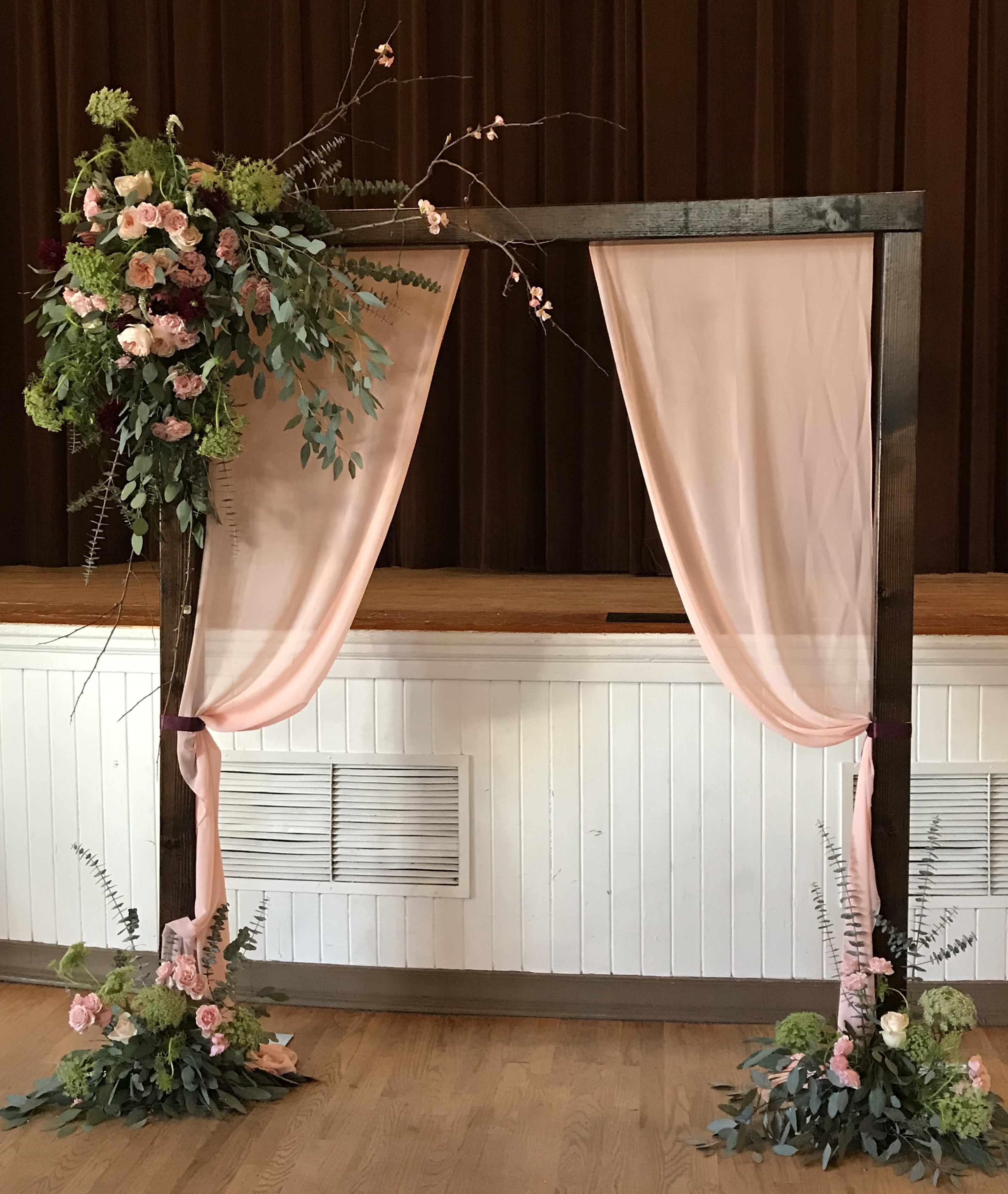 Wooden arbor with bohemian flower types and draping, it has feather eucalyptus, roses, spray roses, dahlias, queen anne's lace, baby blue eucalyptus with blush chiffon drapes. 