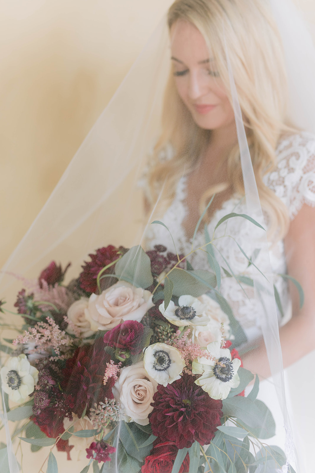 Bride with veil looking at the bouquet of burgundy, blush and ivory