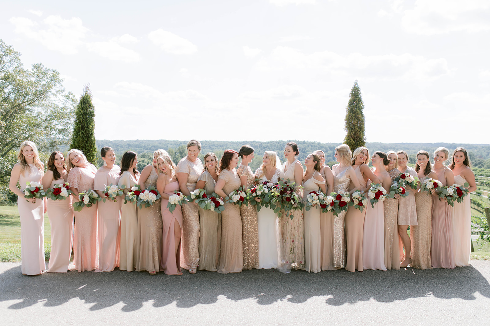 Bride with bridesmaids wearing dresses in a tonal range in blush and gold holding burgundy, blush and ivory bouquets