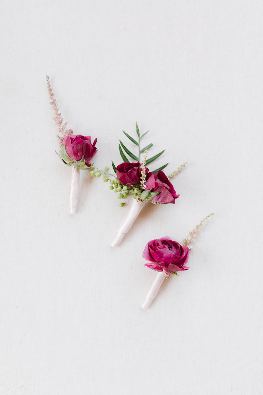 Boutonniere in burgundy rananculus with greenery shown wrapped in blush satin ribbon