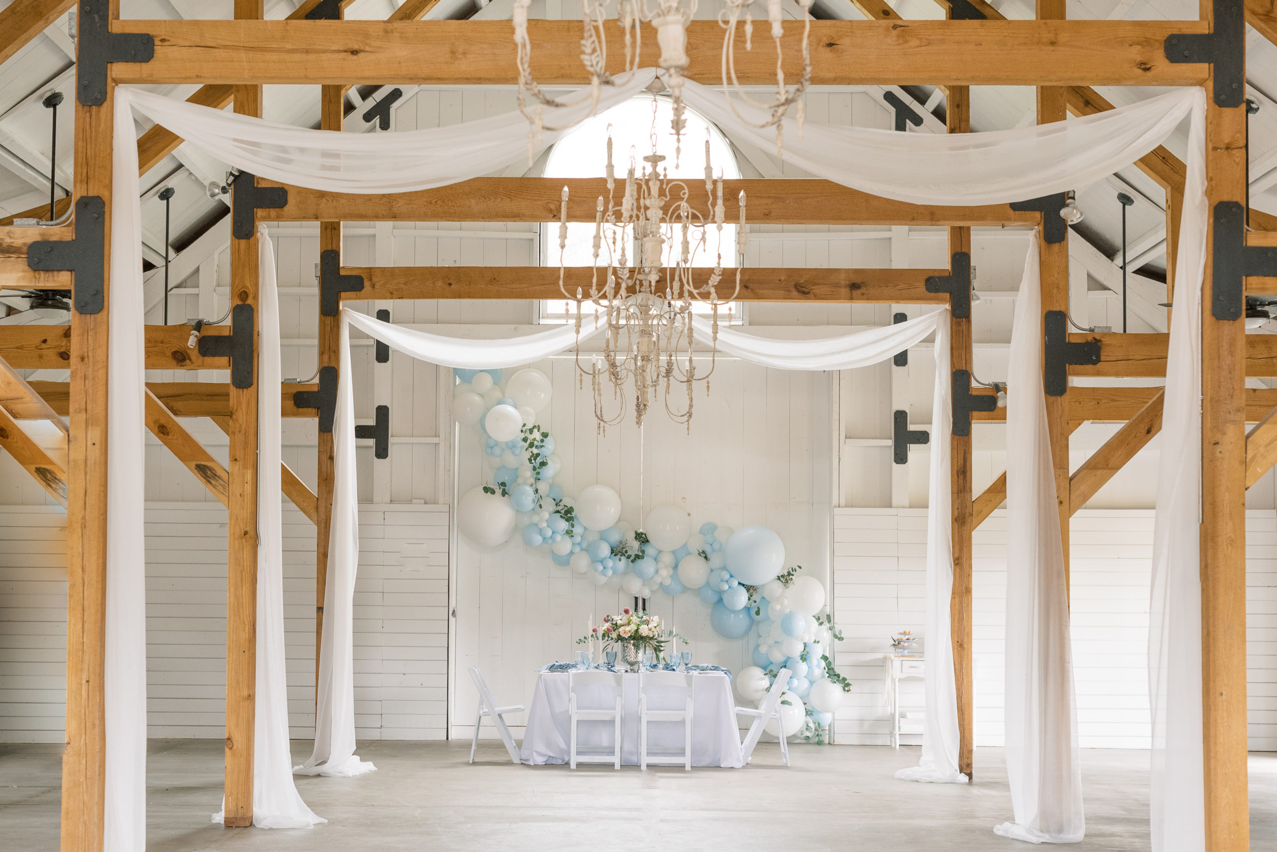 interior photo showing exposed wooden beams, ivory candelabras and white chiffon drapes with balloons in white and pastel blue